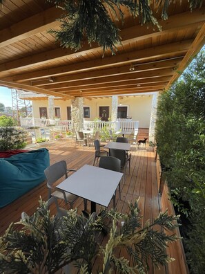 An outdoor patio with wooden flooring, white tables, and chairs. There are potted plants and a dog on the patio.