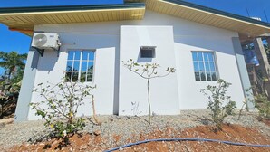 A white building with two windows, a small air conditioning unit, and a tree in front.
