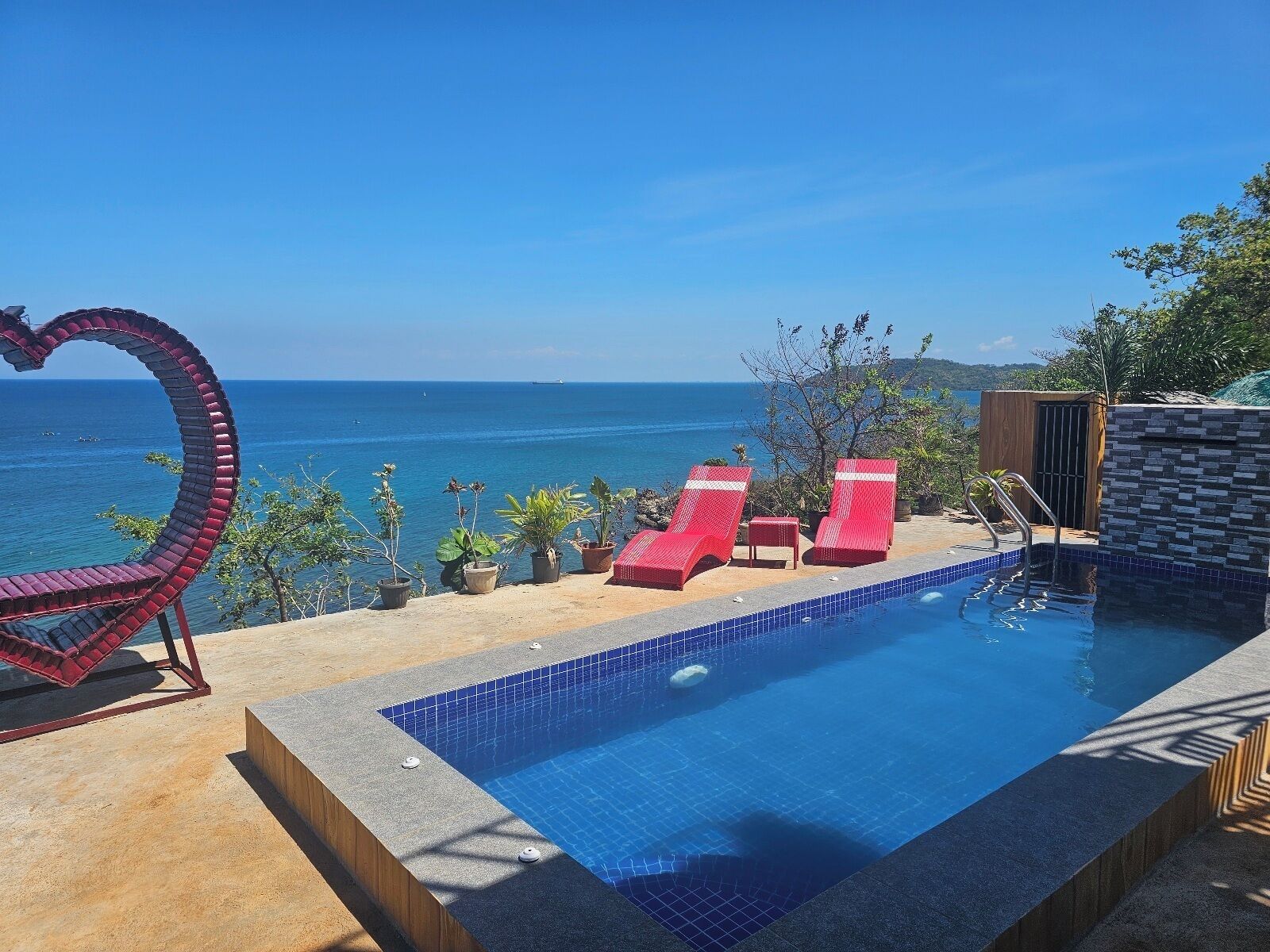 A swimming pool with red lounge chairs and a heart-shaped decoration, overlooking a clear blue sea.