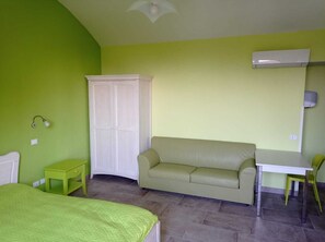 Une pièce comprenant un lit, un canapé vert, une table blanche, une petite table de chevet verte, une armoire blanche et un ventilateur de plafond.
