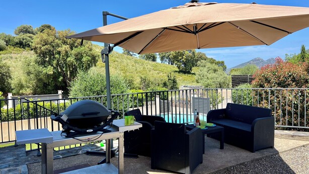 En udendørs terrasse med grill, spisebord og udendørs møbler under en stor parasol.