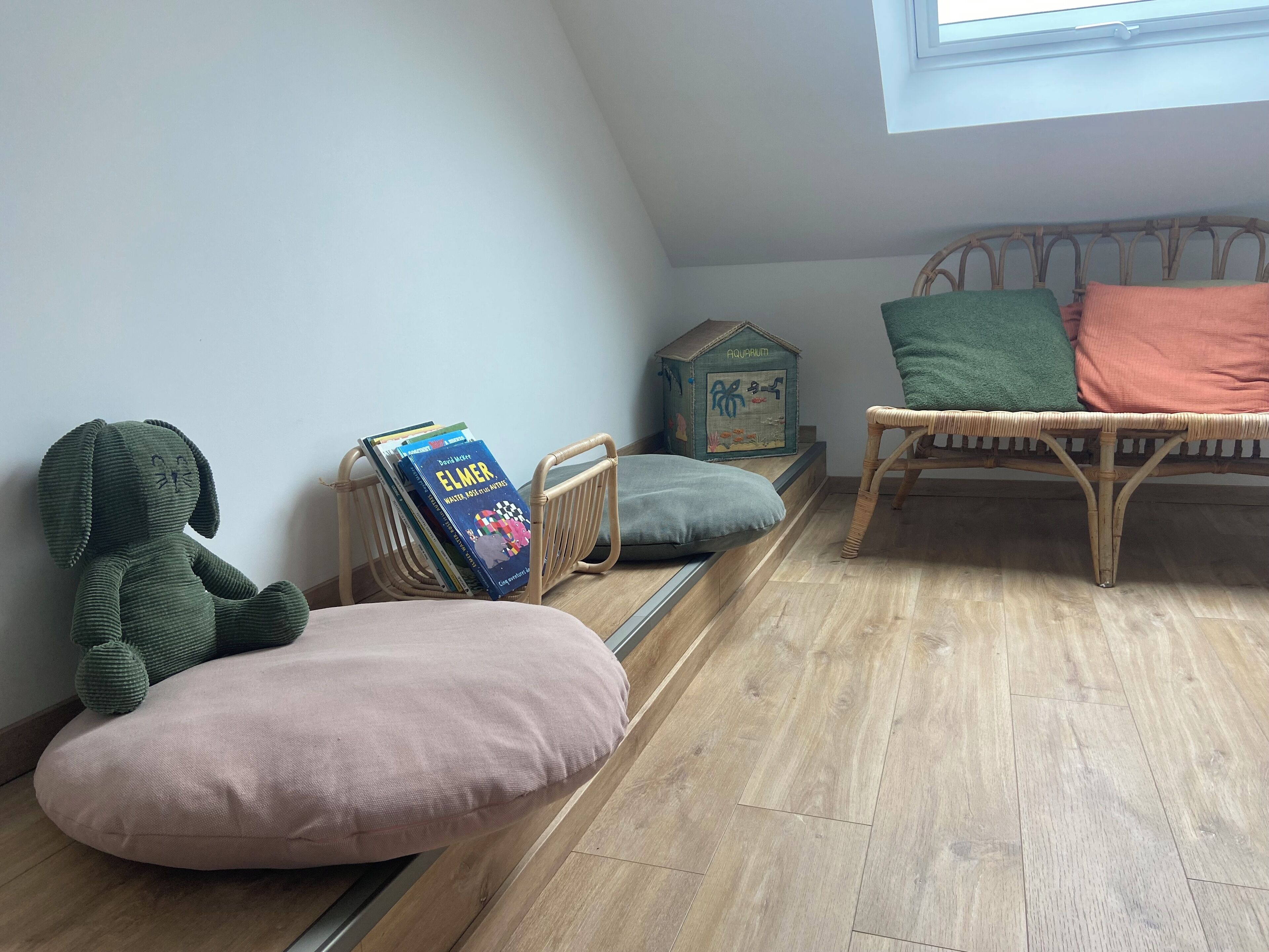 A cozy room with a wooden floor, a green plush toy, a wicker chair with cushions, a small wooden box, and a stack of books.