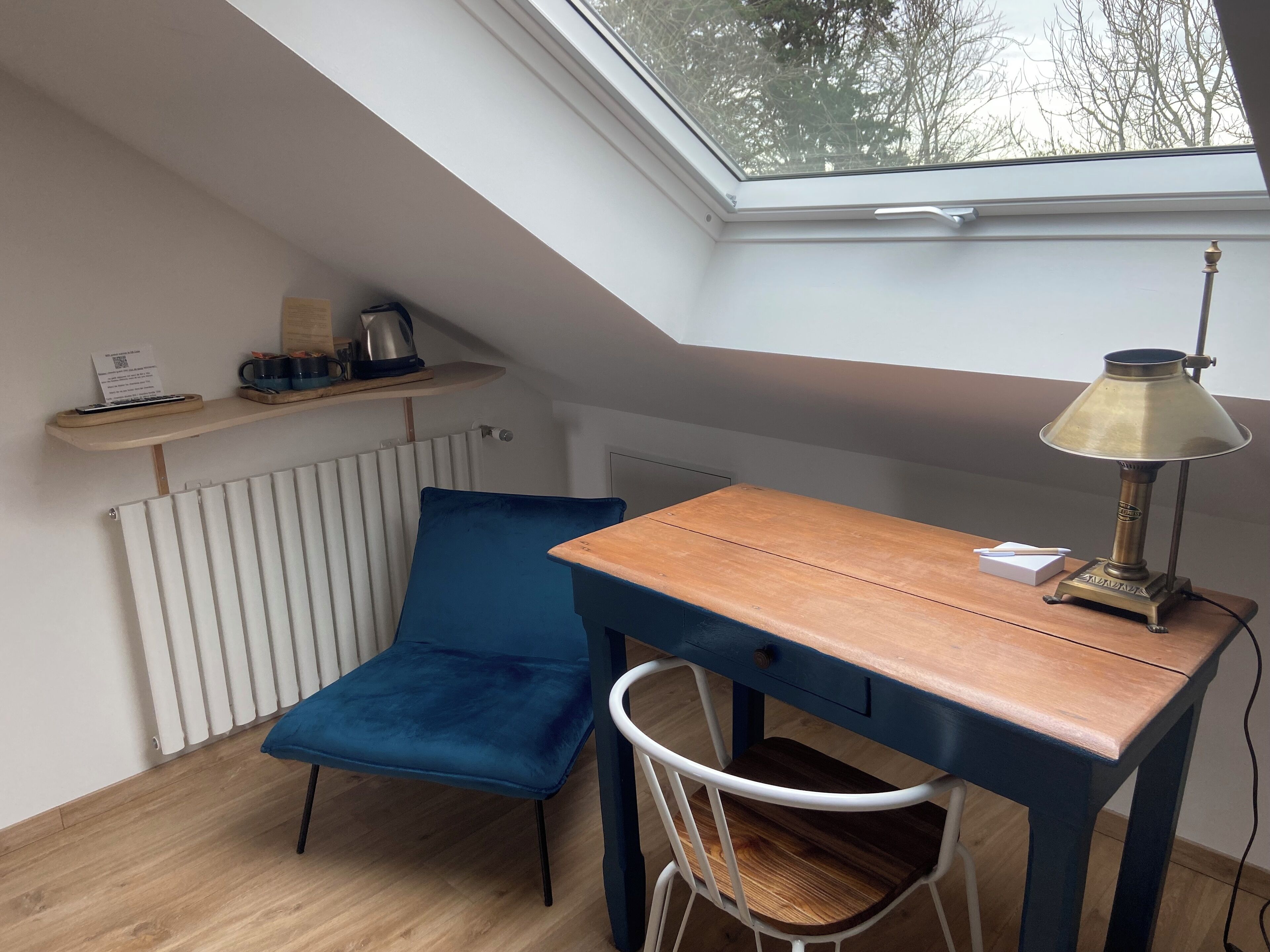 A small, cozy room with a slanted ceiling, a wooden desk, a blue chair, a radiator, and a skylight.