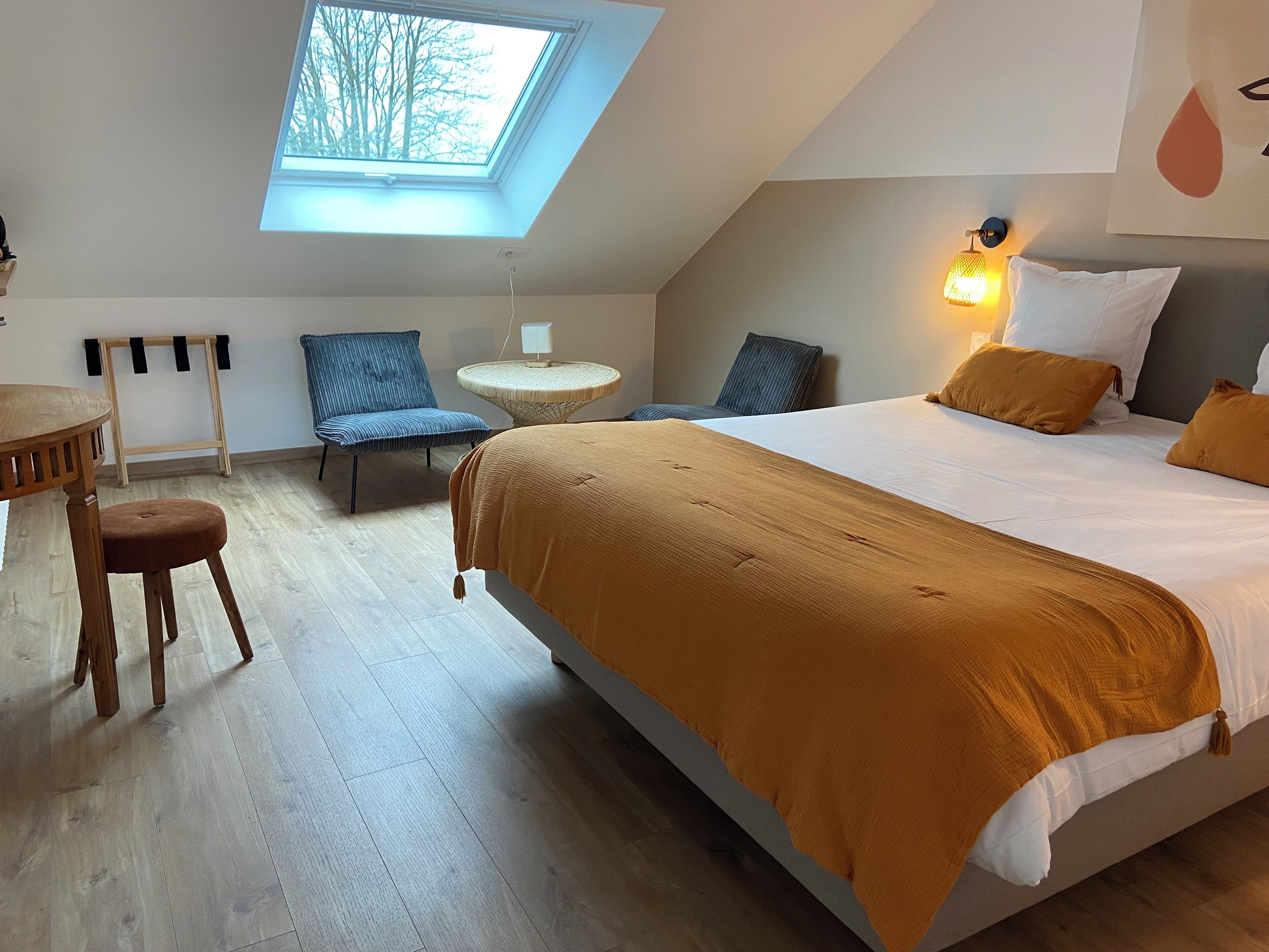 Un dormitorio con tragaluz, una cama con manta marrón, dos sillas, una mesita redonda y una lámpara de pared.