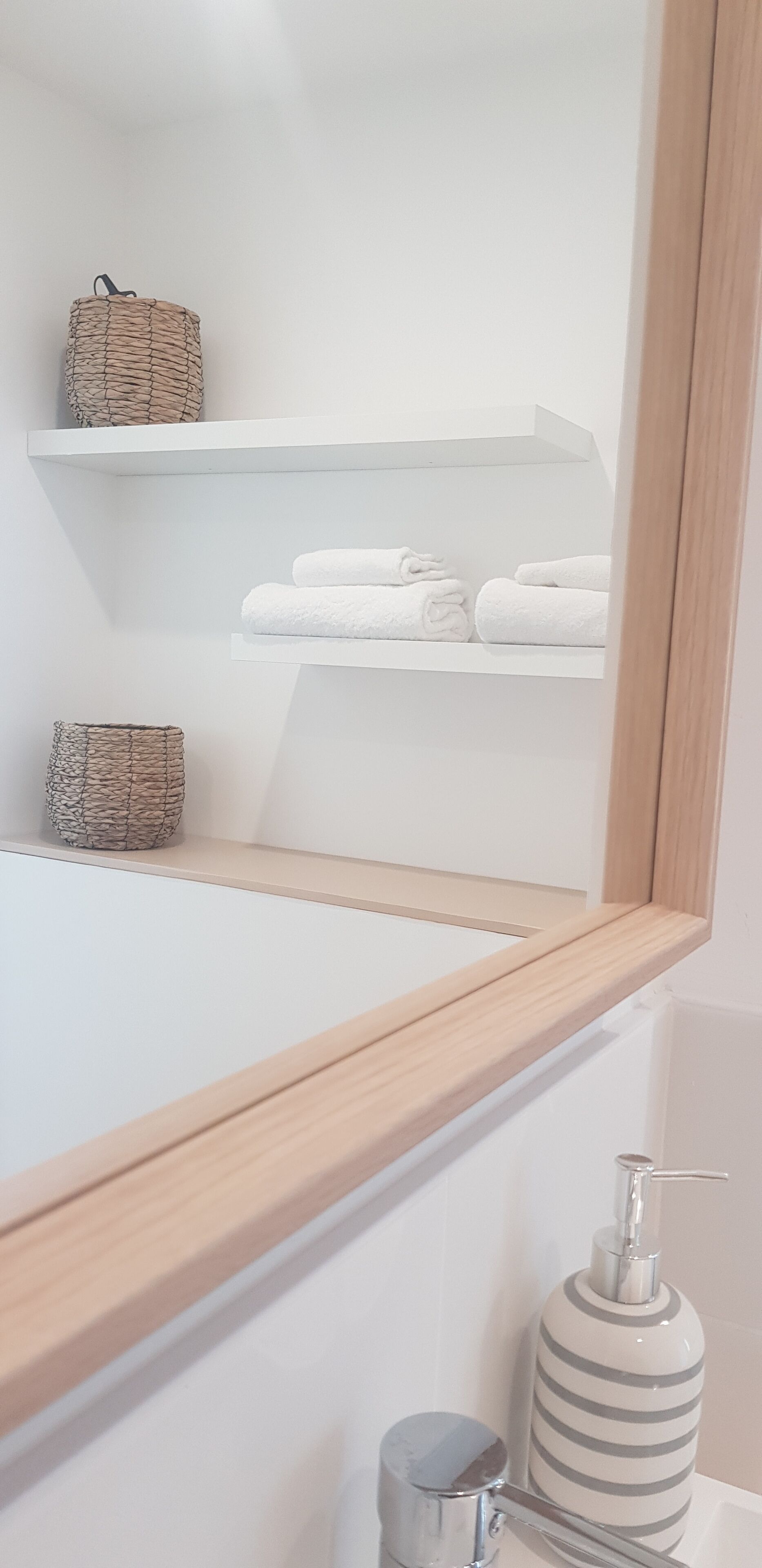 Een badkamer met een houten wastafelmeubel, een spiegel, een handdoekrek met opgevouwen handdoeken en een zeepdispenser.