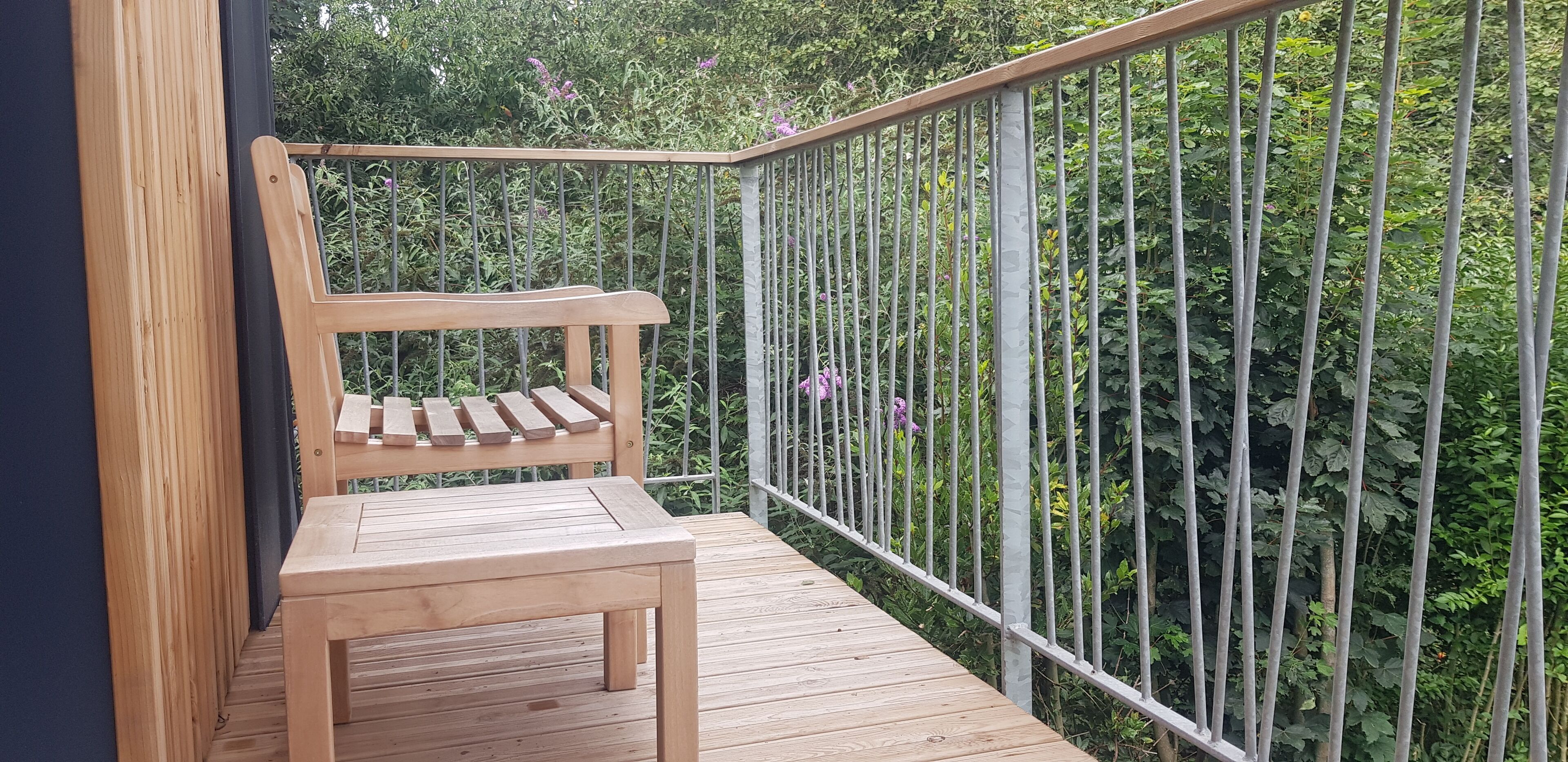 Una terraza de madera con barandilla metálica, un banco de madera y abundante vegetación al fondo.