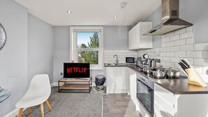 A modern kitchen with white cabinets, a stainless steel range hood, a microwave, and a sink. There's a window with a view of trees, a chair, and a TV displaying the Netflix logo.