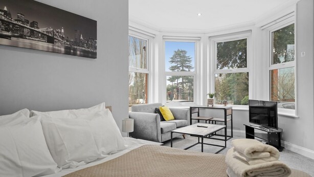 A modern hotel room with a large bed, a grey sofa, a small table, a TV, and large windows offering a city view. - Luxurious Spacious Flat Maidstone with parking (Maidstone)