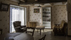 Ein Zimmer mit freiliegenden Holzbalken, einem großen Fenster mit Vorhängen, einem Holztisch, zwei Ledersesseln, einem Bücherregal und einem dekorativen Gemälde.