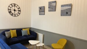 Un salon avec un canapé d’angle, une petite table blanche, un fauteuil jaune et une horloge murale. Il y a trois tableaux accrochés au mur.
