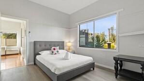 A bedroom with a grey bed, white bedding, a nightstand with a lamp, and a window with a view of trees and a fence.