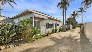 A single-story mobile home with a front porch, surrounded by palm trees and a gravel driveway.