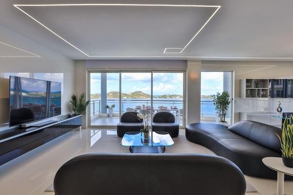 A modern living room with a large TV, comfortable seating, a glass coffee table, and a view of the sea.