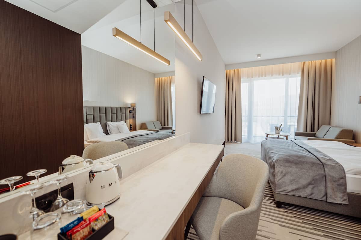 Ein Hotelzimmer mit einem Bett, einem Schreibtisch mit Wasserkocher, einem Sessel, einem Fernseher und einem großen Fenster mit Vorhängen.