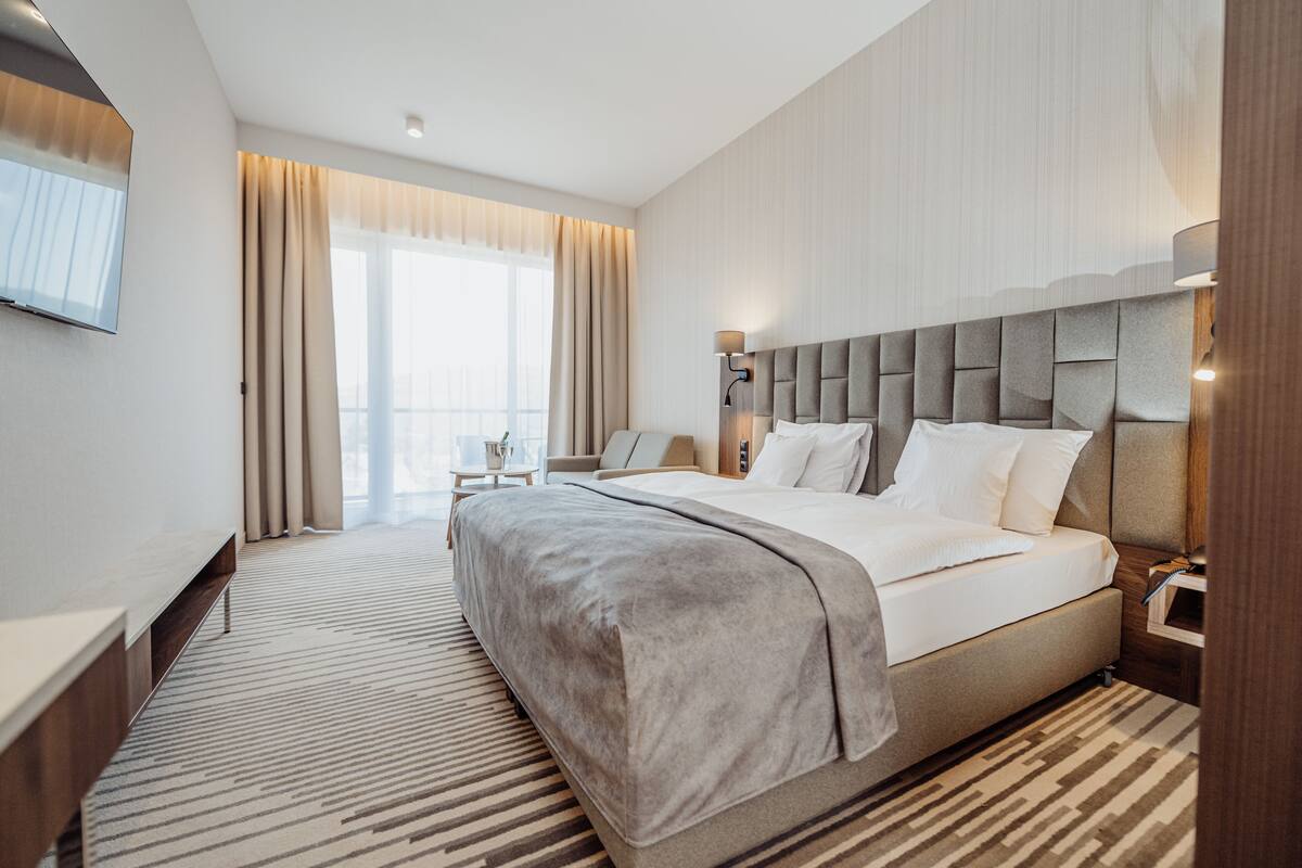 Ein modernes Hotelzimmer mit einem großen Bett, einem Schreibtisch und einem Sessel. Es gibt ein Fenster mit Vorhängen, einen Flachbildfernseher an der Wand und einen Nachttisch mit einer Lampe.