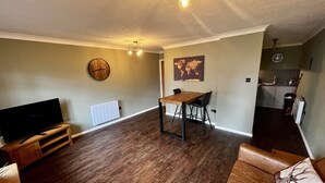 Uma sala de estar moderna com uma mesa de jantar de madeira, um sofá, uma televisão e uma área de cozinha ao fundo.