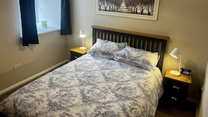 Quarto com cama, duas mesas de cabeceira, um criado-mudo, luminária, quadro e janela com cortinas.