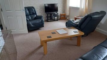 A living room with a wooden coffee table, two black leather sofas, a television on a stand, and a small wooden side table with a teddy bear.