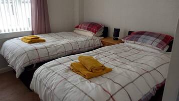 A hotel room with two beds, each with a plaid bedspread and a yellow towel. There is a nightstand with a lamp and a window with curtains.