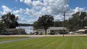 A grassy lawn with a concrete patio, a white fence, and a view of a lake and buildings in the background. - Lake Murray Lake Views Galore (Prosperity)