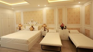 Una habitación de hotel con una cama grande, una cama más pequeña, dos camas individuales, una mesita de noche con una lámpara y una mesita auxiliar con un florero.