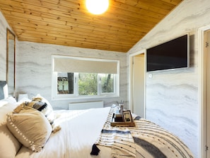 A bedroom with a wooden ceiling, a large window with a view of greenery, a bed with white bedding, and a TV mounted on the wall. - Lovely 3-bed House in Carnforth (Carnforth)