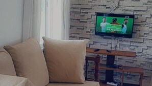 A living room with a beige sofa, a television mounted on a stone wall, and a wooden side table with a remote control. - Stay.Plus Nyandarua Road Apartment Nyali (Mombasa)