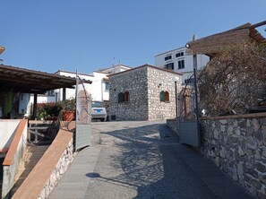 Uma rua estreita com edifícios de pedra, um carro estacionado em frente a uma casa de pedra, e um céu azul límpido.