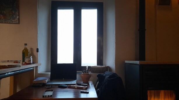 Een kamer met een houten bureau, een laptop, een fles en een pot op het aanrecht. Er is een open haard en een raam met uitzicht.
