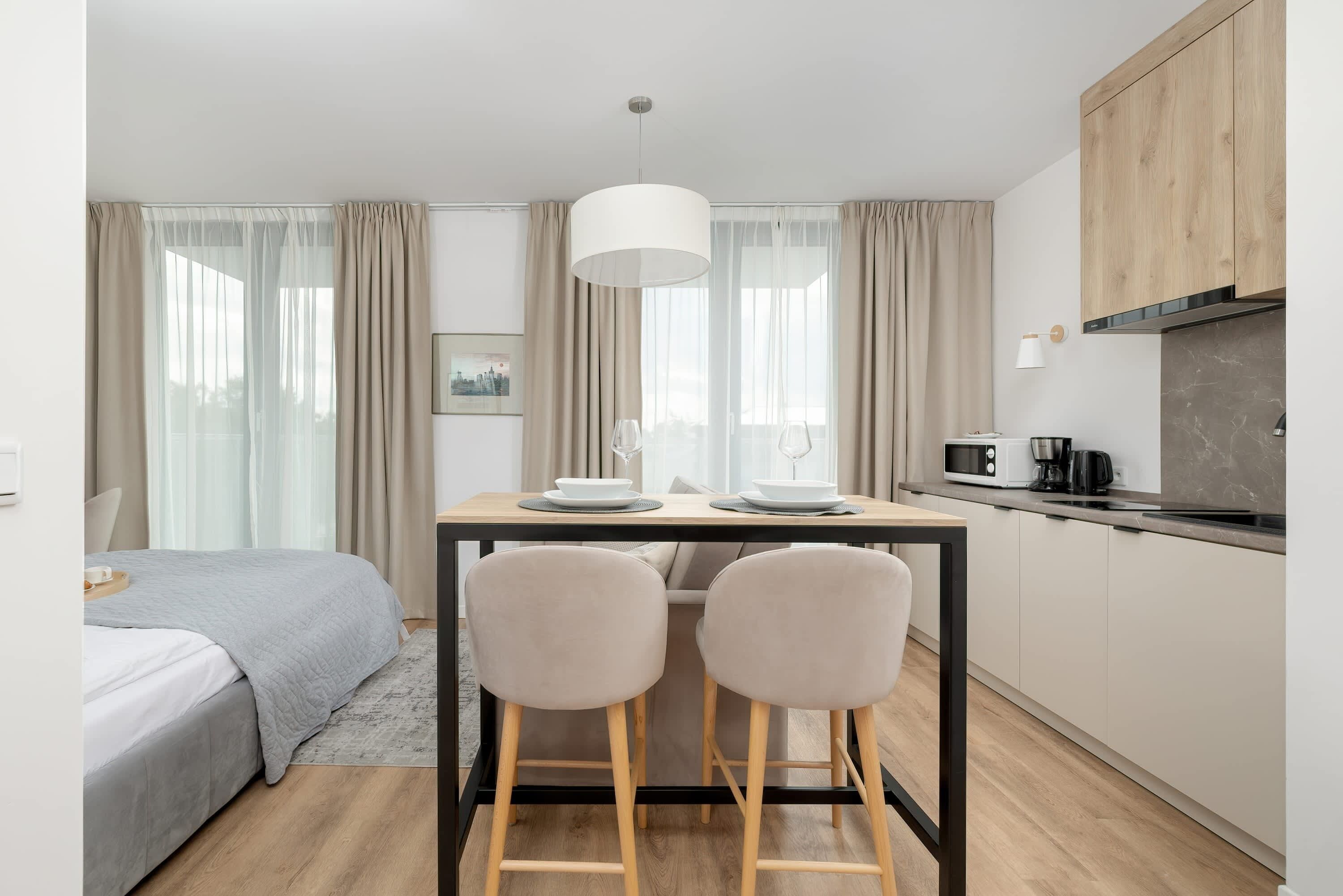 A compact hotel room with a bed, a small dining table with two chairs, a kitchen area with a microwave, and a hanging light fixture.