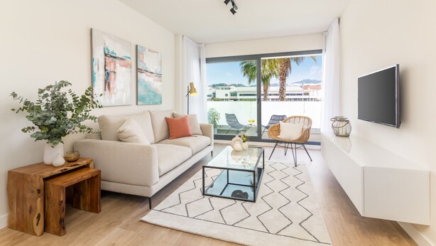 A modern living room with a sofa, coffee table, and a TV mounted on the wall. There is a large window with a view of palm trees and buildings.