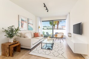 Una sala de estar moderna con un sofá, una mesa de centro y un televisor montado en la pared. Hay un ventanal con vistas a palmeras y edificios.