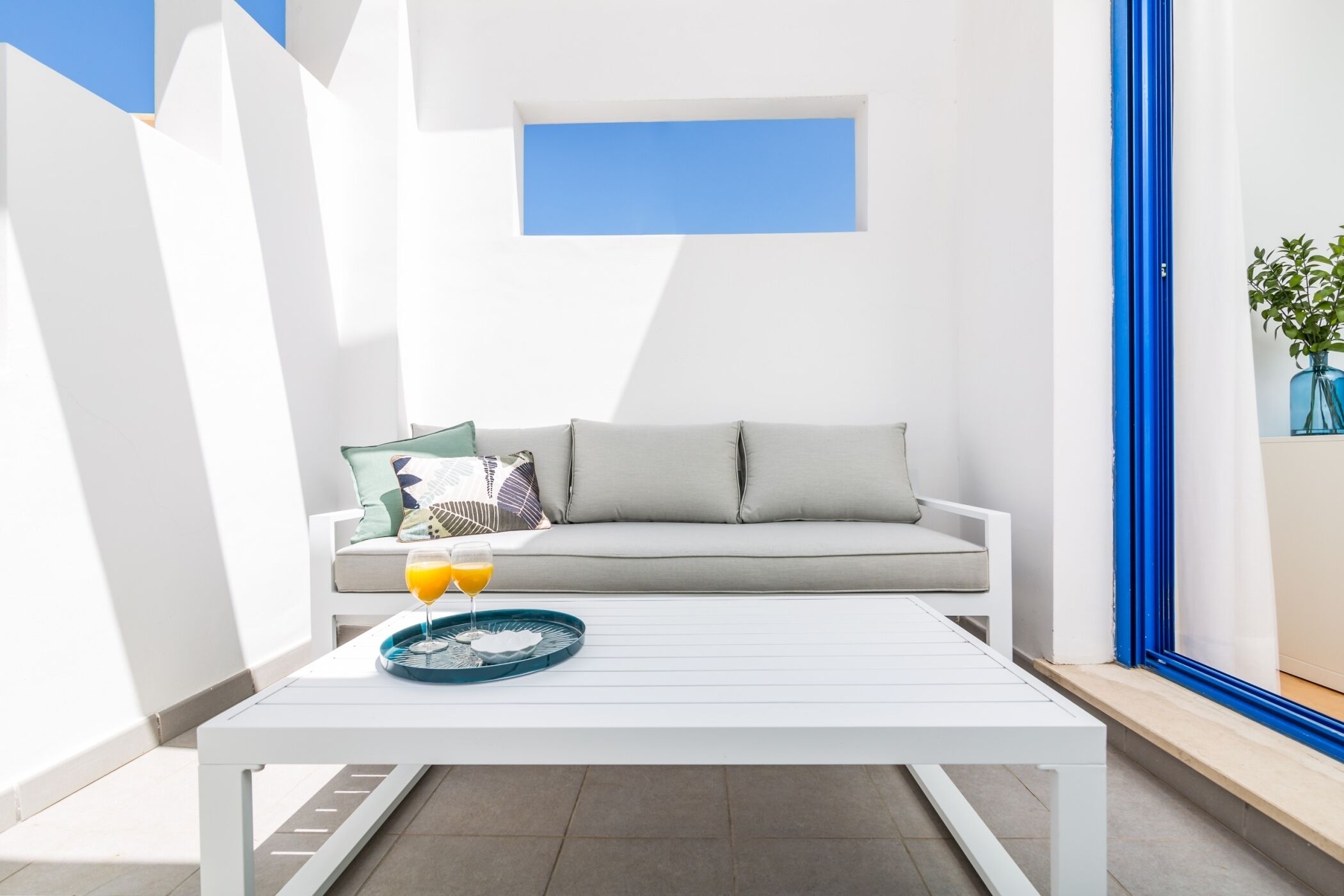 Een moderne buitenruimte met een witte tafel, een bank met kussens en een raam met blauwe lijst.