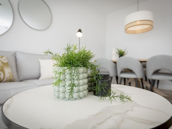 Una sala de estar moderna con una mesa de centro redonda de mármol, un florero de vidrio decorativo con plantas verdes, y un comedor al fondo.