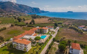 Vista aérea de un complejo turístico costero con varios edificios, una piscina y vistas al mar y a las montañas.