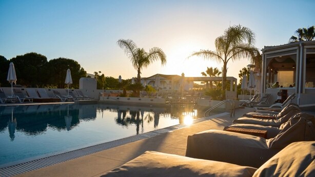 Un área junto a la piscina con sillones de descanso y sombrillas, cielo despejado y palmeras.
