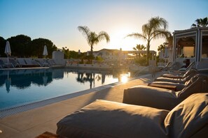 Un área junto a la piscina con sillones de descanso y sombrillas, cielo despejado y palmeras.