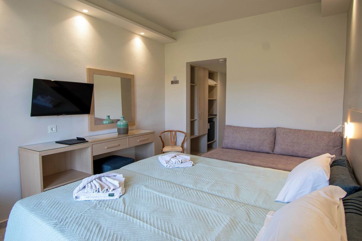 A hotel room with a bed, a desk with a flat-screen TV, a chair, a wardrobe, and a bedside table.