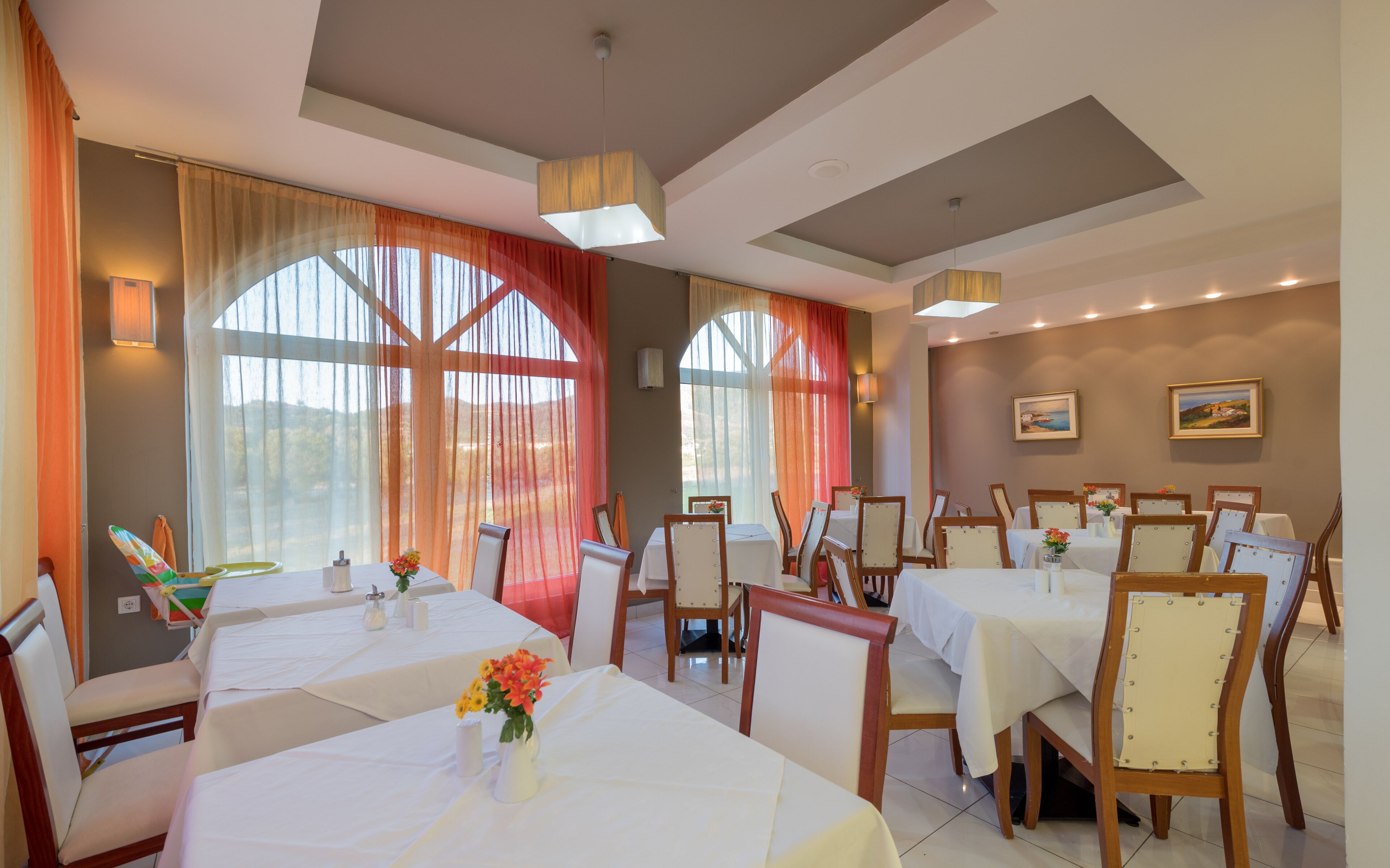 A dining area with tables set for service, large windows with curtains, and artwork on the walls.