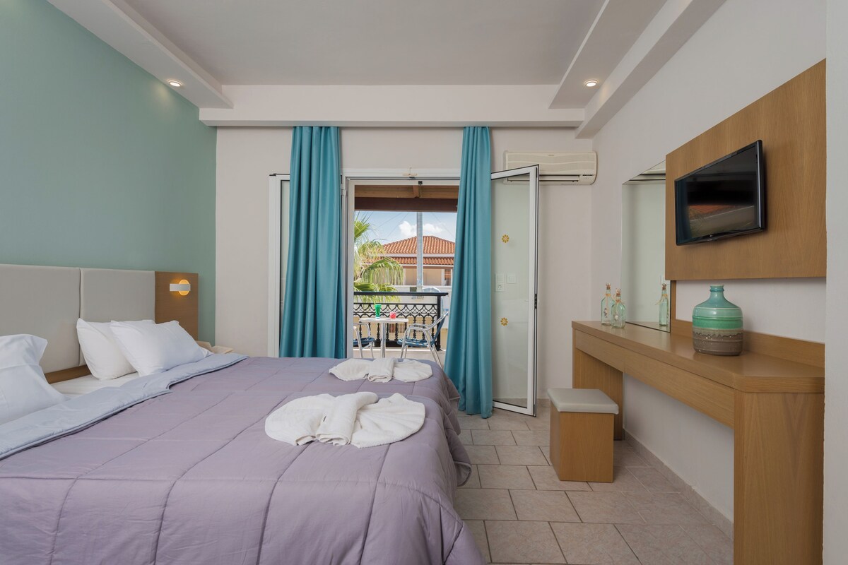 A hotel room with a bed, a TV mounted on the wall, a small table, and a balcony with curtains.