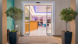 Una entrada de hotel moderna con puerta de cristal, un mostrador de recepción y dos plantas en macetas.