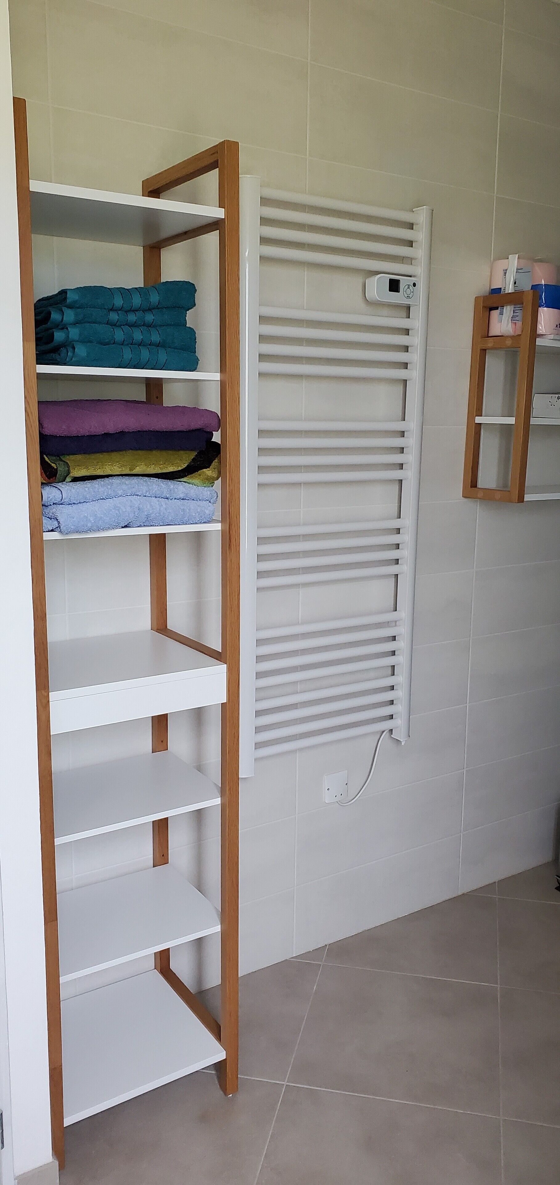 Une salle de bain avec une étagère en bois où sont pliés des serviettes, un radiateur pour serviettes blanc et une petite étagère contenant des articles de toilette.