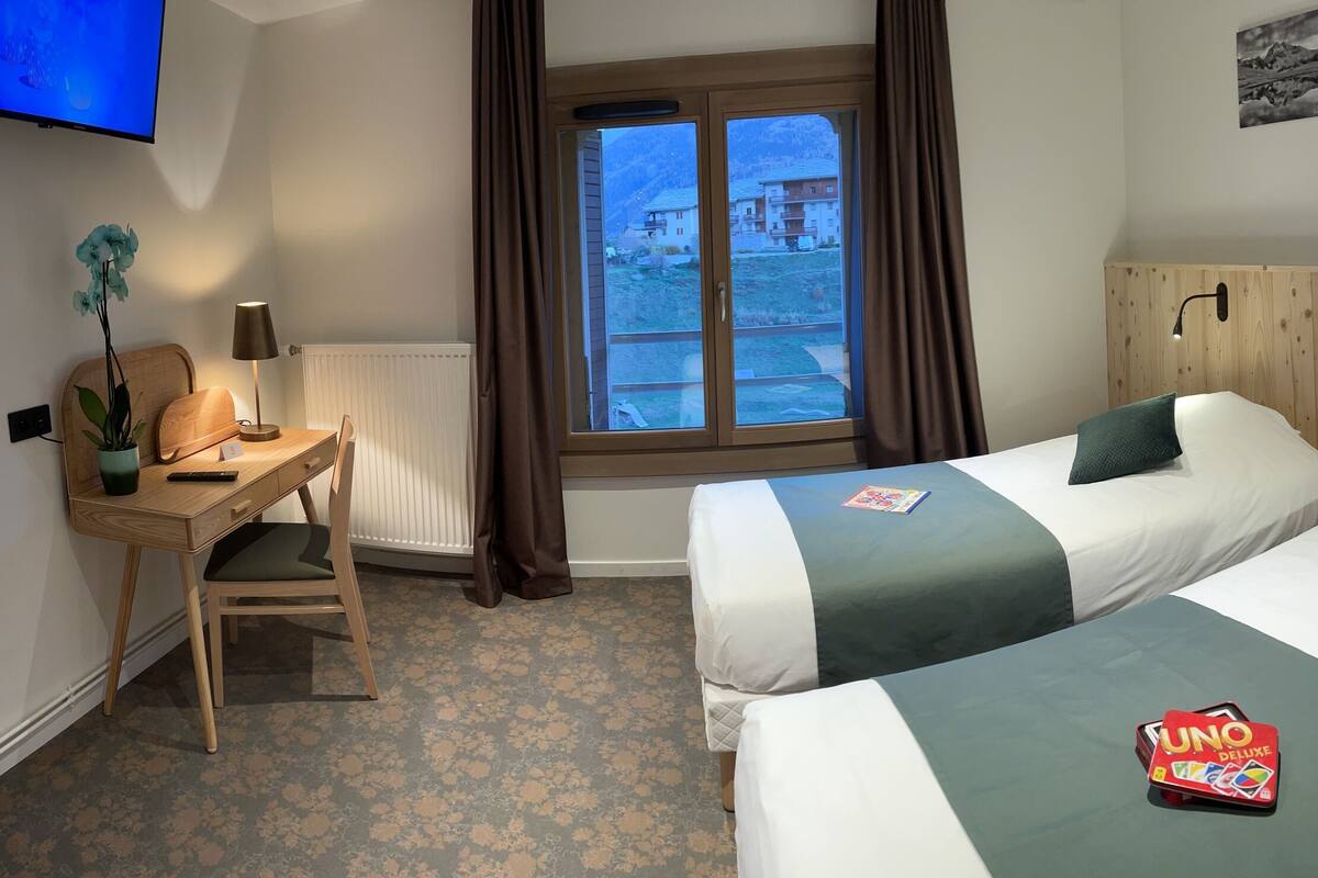 Ein Hotelzimmer mit zwei Betten, einem Schreibtisch, einem Stuhl, einem Fernseher und einem Fenster mit Vorhängen, mit Blick auf Gebäude.