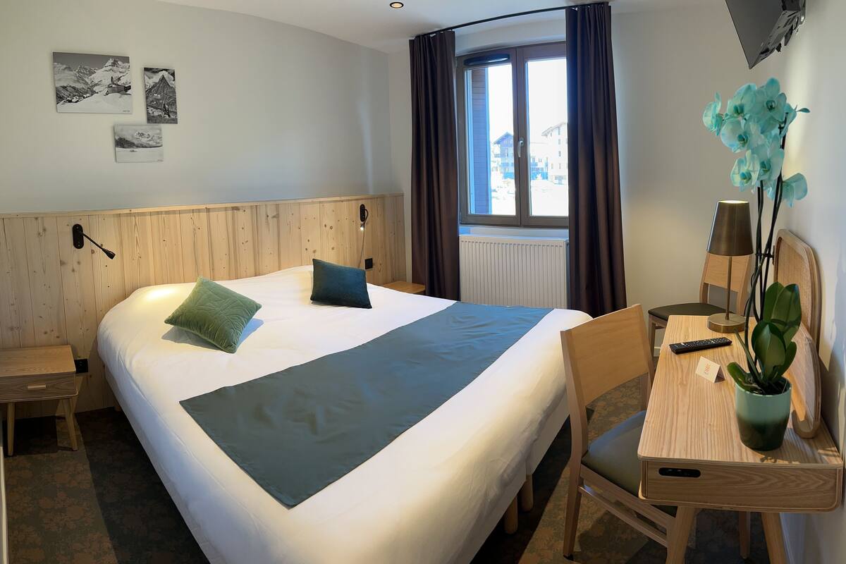 Ein Hotelzimmer mit einem großen Bett, einem hölzernen Kopfteil, einem Schreibtisch mit Stuhl, einer Zimmerpflanze und einem Fenster mit Vorhängen.