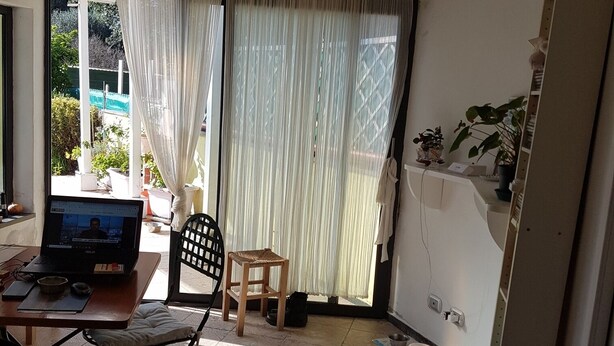 A room with a laptop on a table, a chair, a small stool, a window with sheer curtains, and a potted plant.