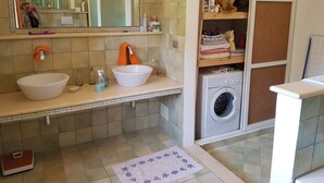 A bathroom with two sinks, a mirror, a shower, a washing machine, and a shelf with folded towels.
