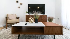 A modern living room with a flat-screen TV mounted on the wall, a wooden coffee table, a beige armchair, and decorative wall art. - Private Chic 2br | 10 min to Downtown| Fast Wifi (Denver)