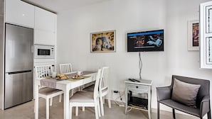 A modern kitchen with white cabinetry, a stainless steel refrigerator, and a microwave. There is a dining table with white chairs and a small TV mounted on the wall.