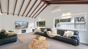A spacious living room with a large TV, leather sofas, a coffee table, and a view of a house through large windows.