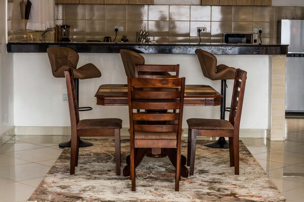 A dining area with a wooden table and four chairs, a kitchen in the background, and a tiled floor.